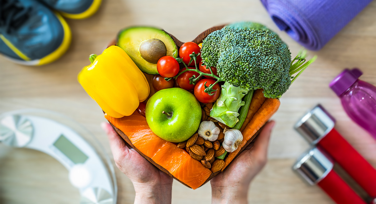 Hands holding a bowl of fresh foods near exercise equipment, a water bottle, and a scale.