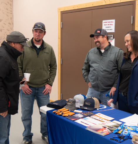 Four attendees visit an exhibitor table displaying hats, informational materials, and promotional items.