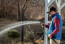 Man blue puffy vest and red black plaid shirt watering in his yard 