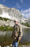 Larry Munn in front of small lake with snowy  granite peak and blue skies behind him 