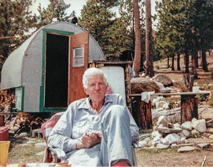White-haired man wearing light blue denim in camp chair with silver sheep wagon and ponderosa pine trees in the background 