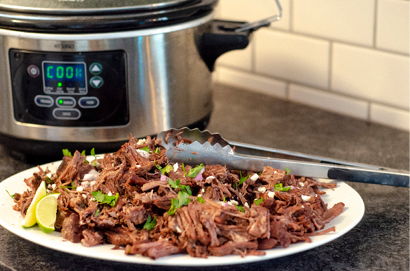 Pulled pork on white plate with cilantro  red onion and lime wedges for garnish  Silver slow cooker in the background 