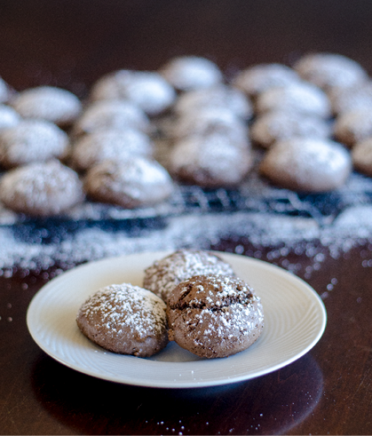 3 round chocolate cookies dusted with white powdered sugar on a plate  Several cookies on a cooling rack in the background  