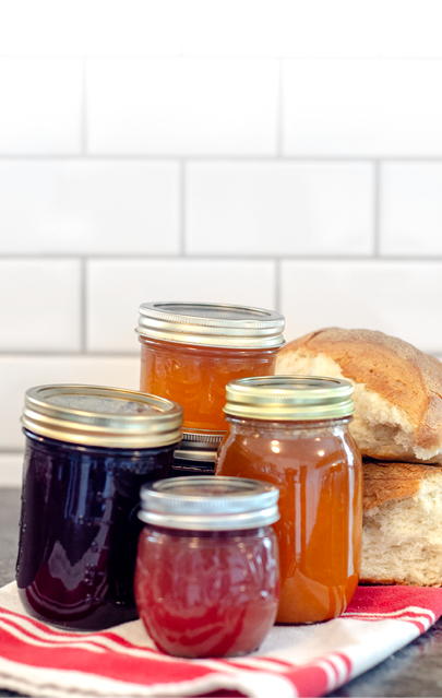 Five glass jars of homemade jams with torn loaves of rustic white bread rest on a red and white towel on a kitchen counter 