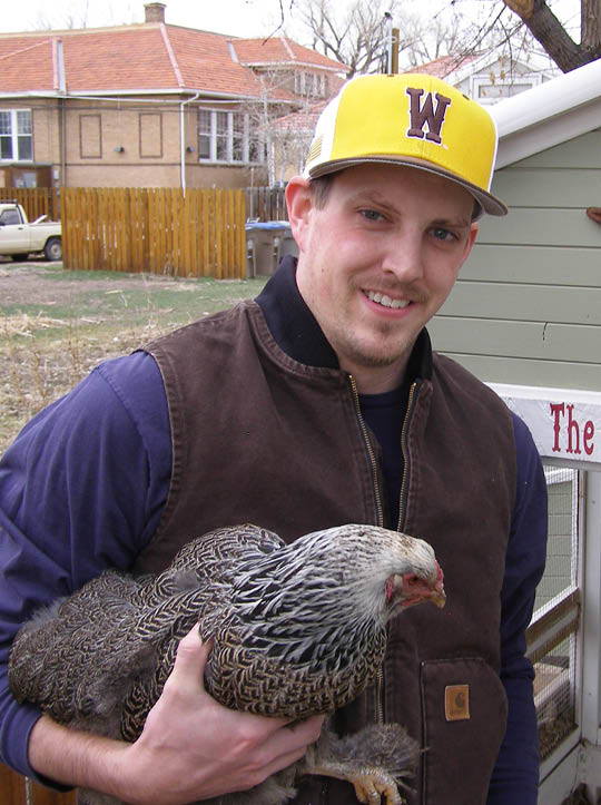 Man in brown vest, blue shirt, brown and yellow baseball cap standing in his backyard holding a black and white chicken