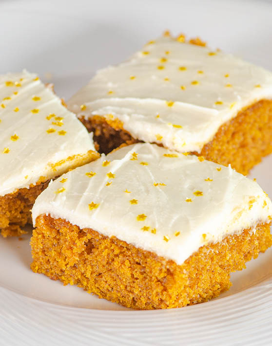Three browned butter frosted pumpkin bars with gold star sprinkles placed on a white plate. 
