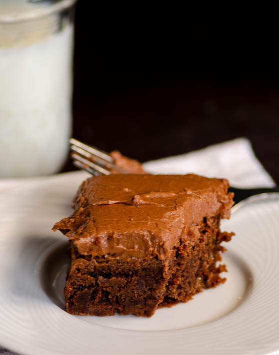 A single frosted brownie on a white plate with a fork in the background. 