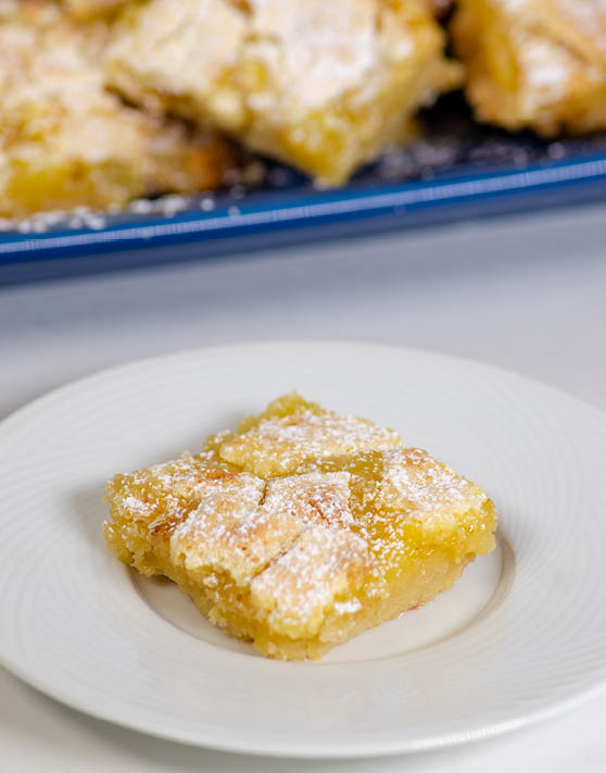 A single lemon bar on a white plate with a pan of lemon bars in the background. 