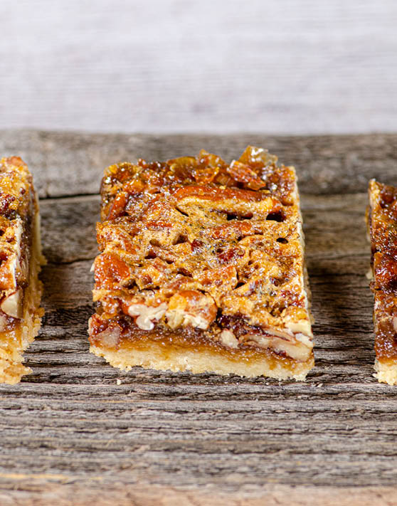 A southern pecan pie bar placed on a wooden surface, with two other southern pecan pie bars in the frame. 