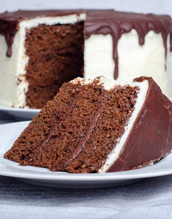 A slice of chocolate cake placed on a white plate, with an entire chocolate cake in the background. 