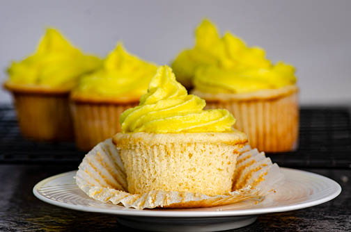 A white cake cupcake with yellow frosting placed on a white plate, with white cake cupcakes in the background. 