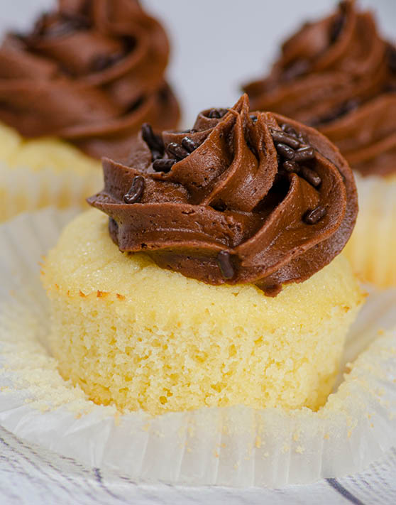 A close-up of yellow cake cupcake with brown frosting, and other cupcakes in the background. 