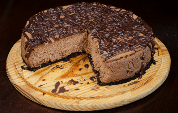 A chocolate cheesecake with choclate frosting drizzled on top and a piece missing from the center, placed on a wooden board. 