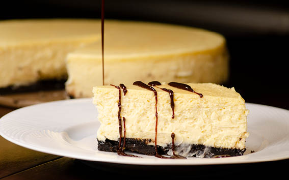 A piece of New York style vanilla cheesecake with a chocolate base, placed on a white plate, and a cheesecake with a piece missing in the background. 