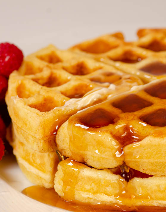 Two ultimate buttermile waffles with syrup drizzled on top and raspberries on the side. 