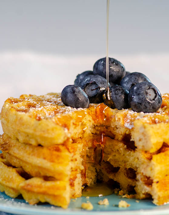 A stack of oat cornmeal waffles with syrup drizzled and blueberries on the top. 