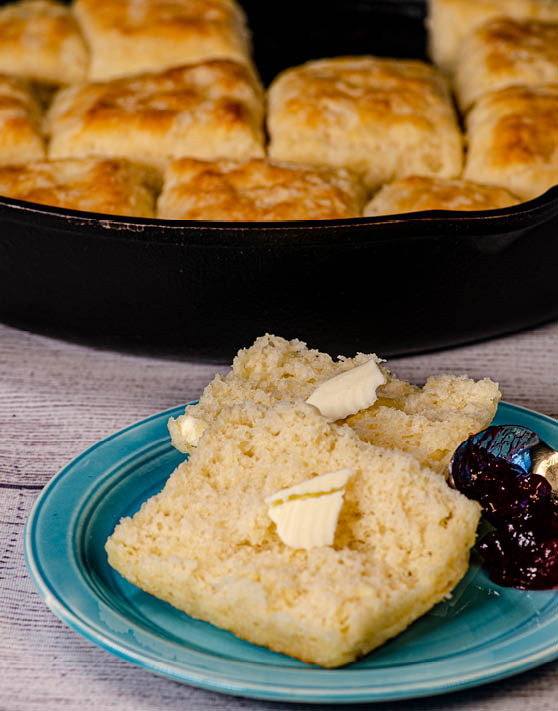 An easy buttermilk biscuit cut in half with butter on both halves and a pan of biscuits in the background. 
