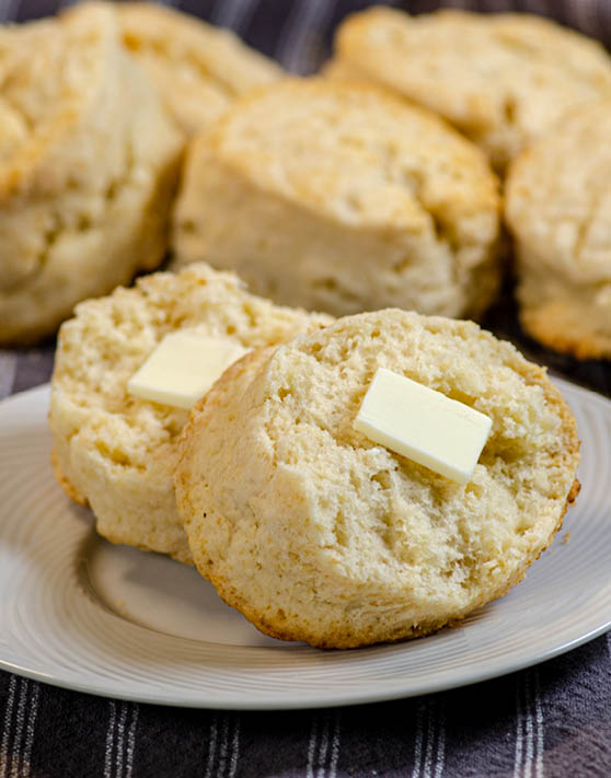 A buttermilk cream cheese biscuit, placed on a white plate, and cut in half with butter and biscuits in the background. 