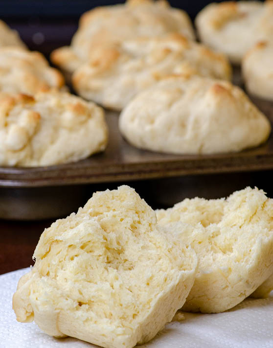 A muffin tin batter roll split in half with a pan of muffins in the background. 