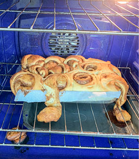 A pan of overflowing cinnamon rolls, with cinnamon roll substance on the bottom of the oven and on a second pan. 