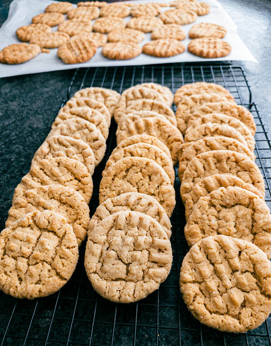 Three rows of classic peanut butter cookies placed on a cooling rack and peanut butter cookies in the background on parchment paper. 