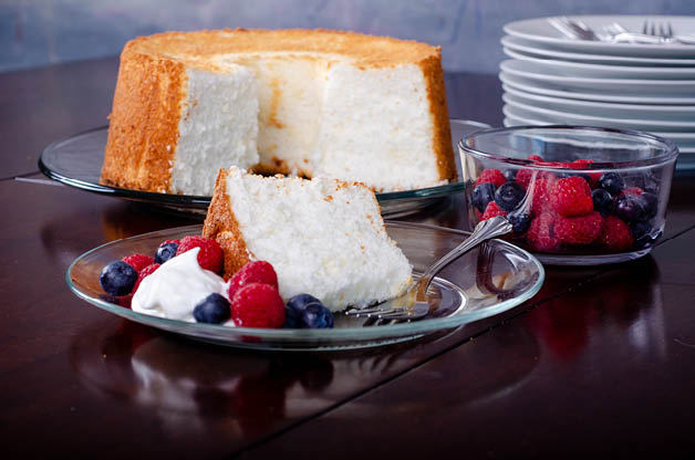 A slice of angel food cake placed on a clear plate with raspberries and blueberries. An angel cake in the background. 