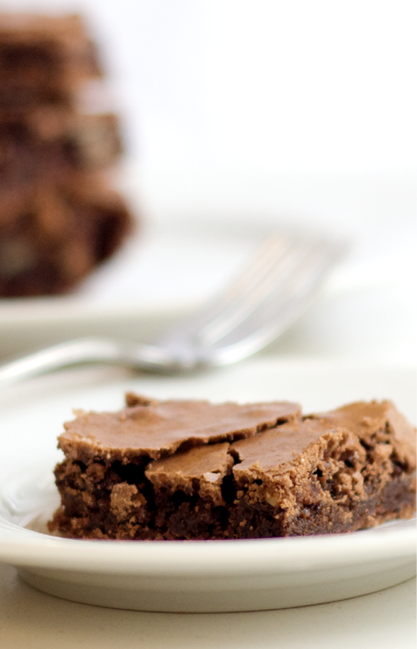 One fudge brownie sits on a white plate with a fork and a stack of brownies in the background 