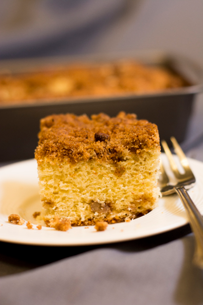 Piece of cinnamon-nut coffee cake on a white plate with a fork  The pan is in the backround 