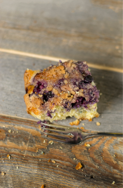 A piece of blueberry coffee cake on a reclaimed wood table with a fork near by