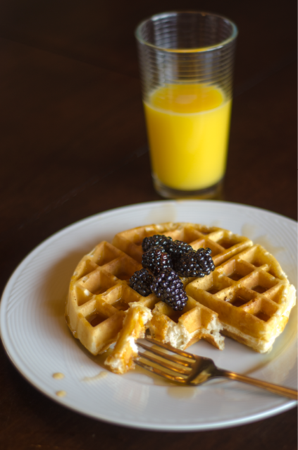 A Belgian waffle missing a few bites with maple syrup and fresh blackberries on a white plate  A large glass of orange juice is in the background 