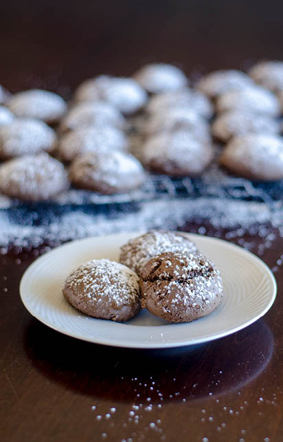 Three chocolate drop cookies dusted with powdered sugar are on a white plate  In the background is a cooling rack with several more of the chocolate drop cookies 