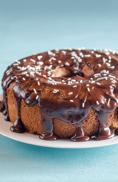 Chocolate cake was baked in a tube pan  It was heavily drizzled with a thick chocolate glaze and then white sprinkles were scattered over the top  The cake is resting on a white plate on an aqua-colored tablecloth 