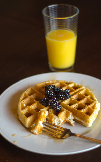 A Belgian waffle missing a few bites with maple syrup and fresh blackberries on a white plate  A large glass of orange juice is in the background 