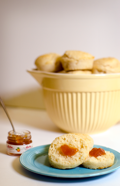 Retro yellow bowl of plain muffins sits in the backround with a blue plate with two muffins  one with strawberry jam  in the foreground 