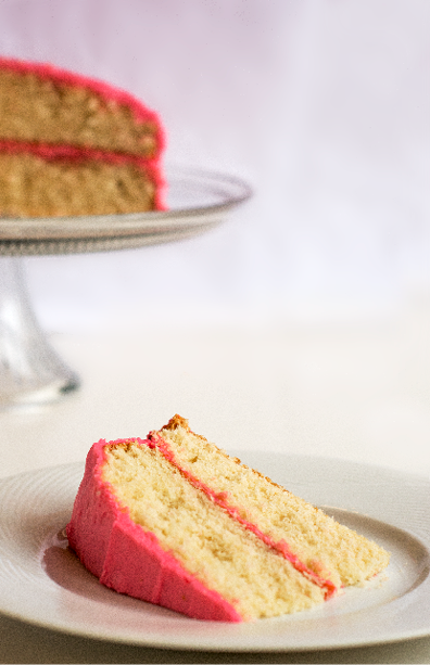 White layer cake with pink frosting  One slice is on a white plate and in the background is the remainder of the cake on a glass cake pedestal 
