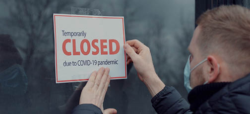 Person in blue medical mask taping sign to glass door  Sign reads,  Temporarily closed due to COVID-19 pandemic  