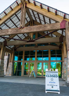Building entrance with large wood beams, glass doors, and an A-frame display board reading,  Visitor Center Closed  