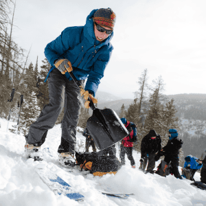 Person wearing a blue jacket and gloves, shoveling snow during a winter outdoor activity, with a group of people in the background on a snowy mountainside.