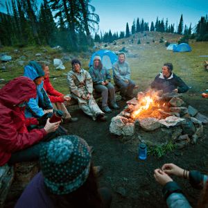 Group of campers sitting around a campfire at dusk, dressed in outdoor gear with tents in the background in a forest clearing