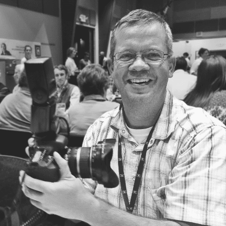 Brad Christensen: A black-and-white photo of Brad Christensen smiling while holding a professional camera, taken at an event with people in the background.