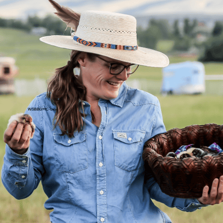 Melissa Hemkin: Melissa Hemkin outdoors, wearing a straw hat with a colorful band, holding a basket and a small chick, showing her involvement in agriculture or animal care.