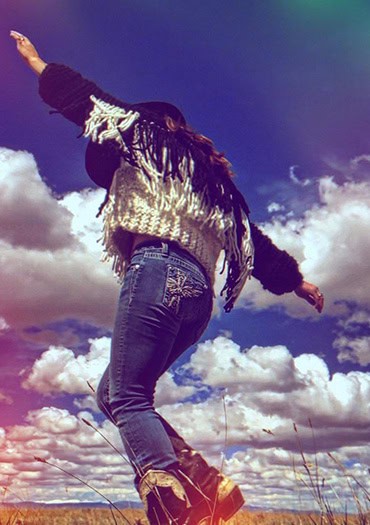 A person wearing blue jeans, platform boots, and a fringed wool sweater dances or balances playfully in a grassy field under a dramatic sky filled with puffy clouds. The image is taken from a low angle, capturing motion and energy, with a colorful lens flare effect adding a dreamy atmosphere.