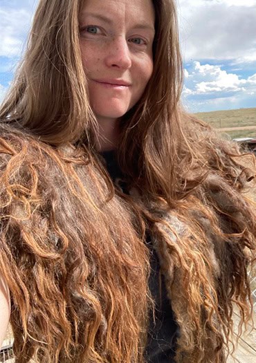 A woman with long, wavy auburn hair stands outdoors under a partly cloudy sky, wearing a vest or garment made from naturally colored, textured wool. She smiles softly at the camera, with open grassland and a wire fence in the background.