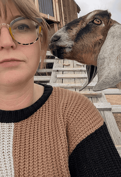 Woman wearing glasses and a brown, black, and white knit sweater sitting on wooden steps, with a brown goat standing behind her and looking toward her face.
