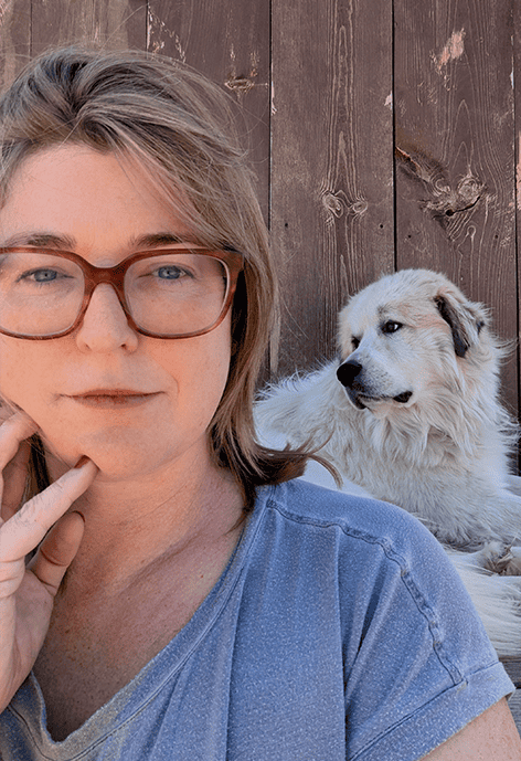 Woman wearing glasses and a brown, black, and sitting on wooden steps, with a white dog sitting behind her and looking toward her face.