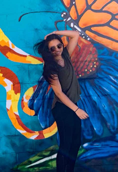 Young woman posing in front of a colorful mural featuring a large monarch butterfly and vivid flower petals. She is wearing sunglasses and a dark outfit, with one hand raised above her head.