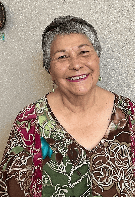 Smiling older woman with short gray hair wearing a multicolored floral blouse, standing against a textured beige wall.