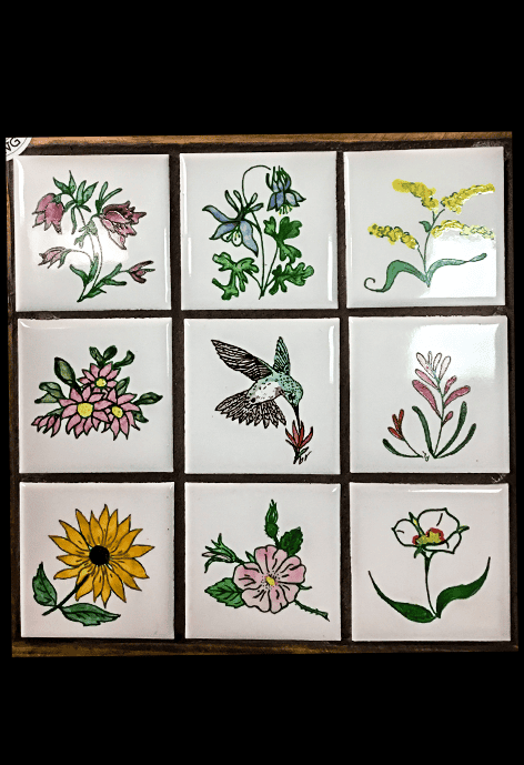 A framed ceramic tile set with nine small squares, each hand-painted with a different botanical or bird design, including wildflowers, a sunflower, and a hummingbird, labeled with a “Made in Wyoming” sticker.