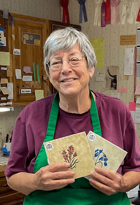 Smiling older woman with short gray hair and glasses wearing a maroon shirt and green apron, holding two decorative tiles with floral artwork in a cozy studio filled with ribbons and notes