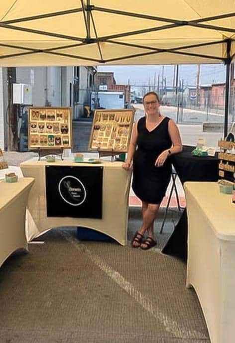 A jewelry artist stands under a cream-colored canopy tent at an outdoor market, surrounded by neatly arranged tables and displays of handcrafted earrings. The artist, wearing a black dress and sandals, smiles beside a sign that reads “S Brown Creations.” Industrial buildings and a clear sky form the backdrop.